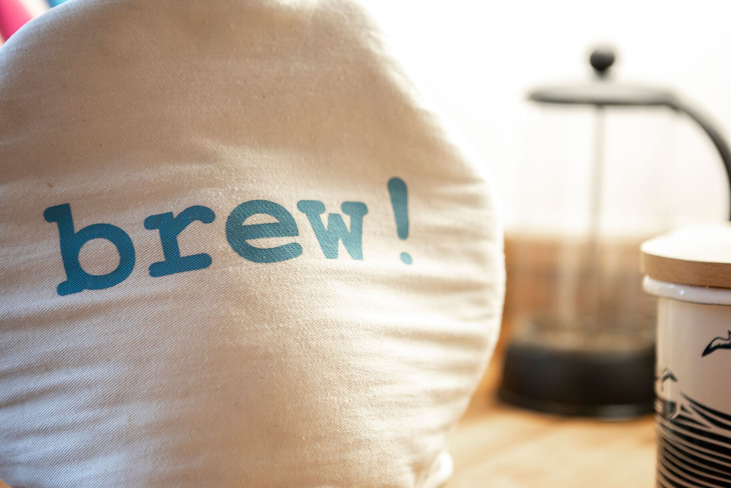 A close-up of a pillow with the word "brew!" on it, a coffee maker, and a cup with a lid.