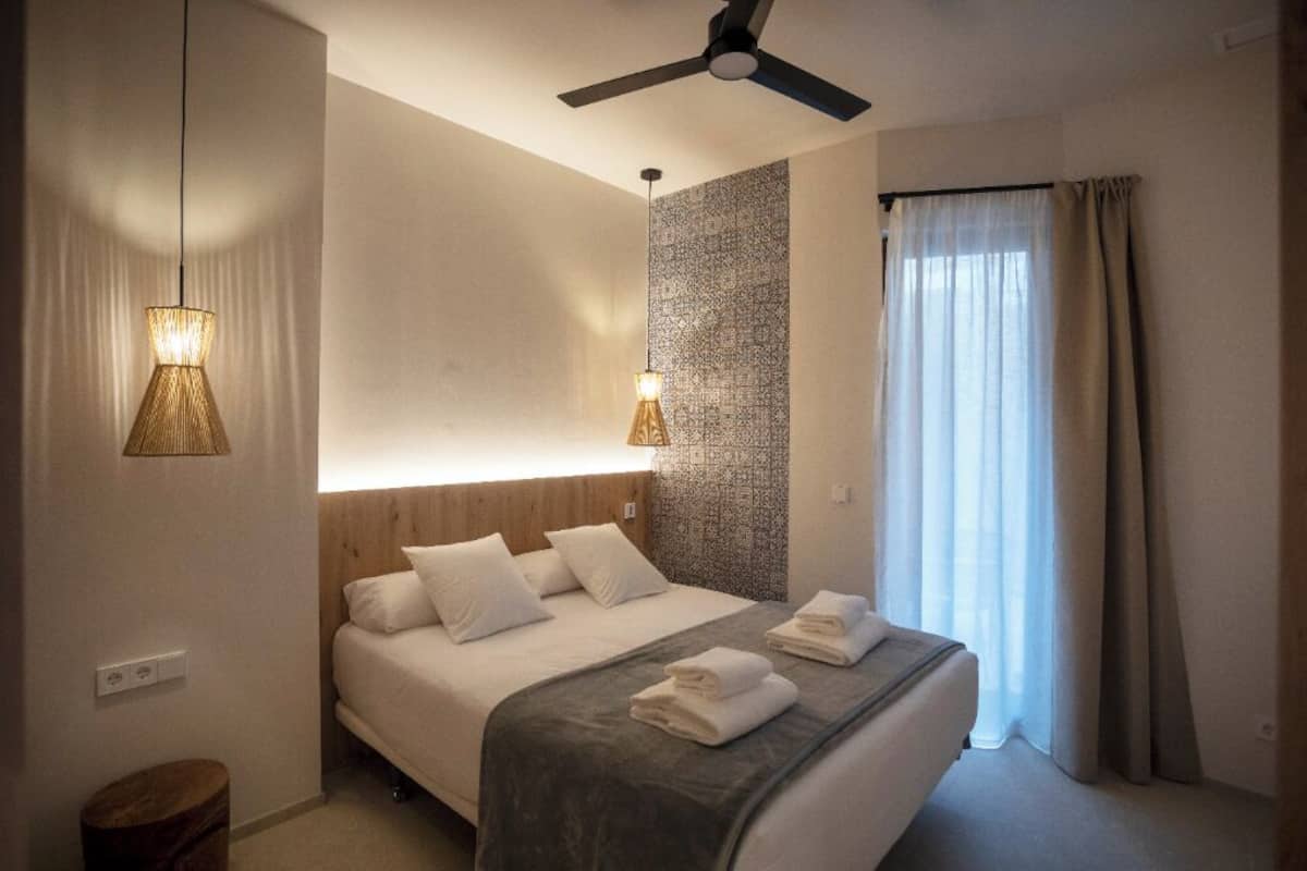 A hotel room with a bed, two bedside lamps, a ceiling fan, and a window with curtains.