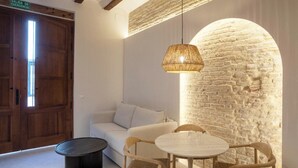 A cozy room with a white sofa, a round wooden table, and wicker chairs. There's a stone wall and a wooden door labeled "SALIDA DE EMERGENCIA". - Mon Suites San Pedro (Valencia)