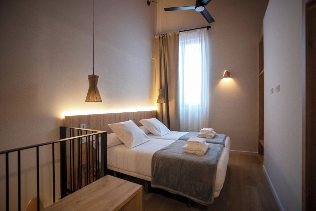 Een slaapkamer met twee bedden, een plafondventilator, een hanglamp en een raam met gordijnen.