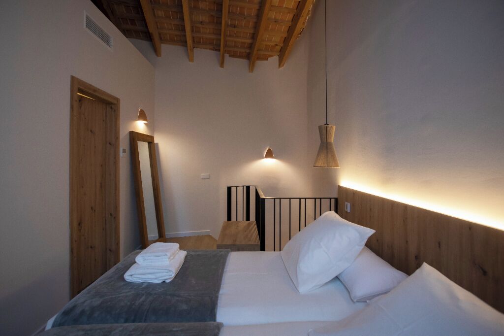 Un dormitorio con techo de madera, una cama con ropa de cama blanca, una mesita de noche con lámpara y un espejo en la pared.