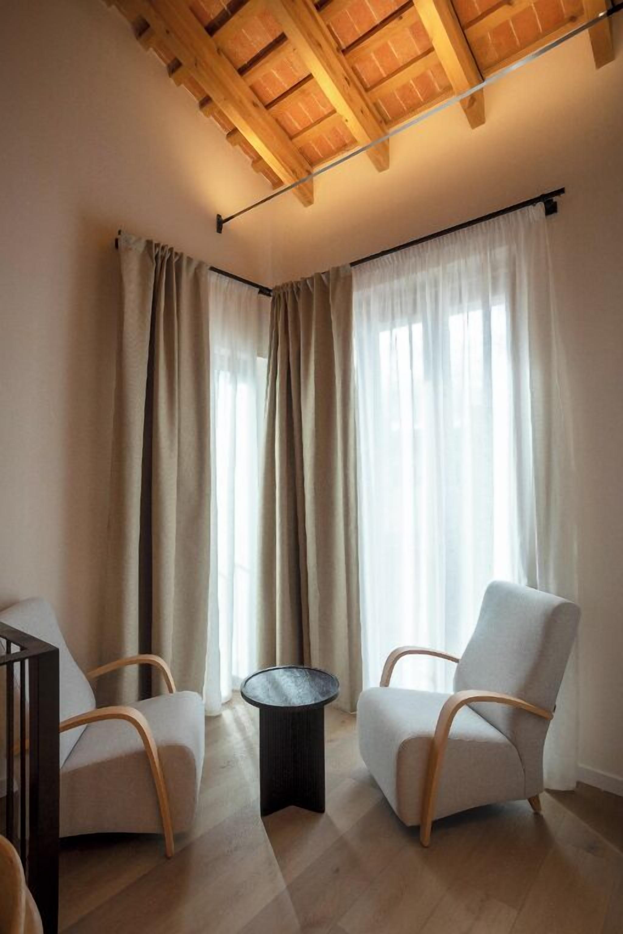 A modern living room with a wooden ceiling, two white armchairs, a small round table, and large windows with sheer curtains.