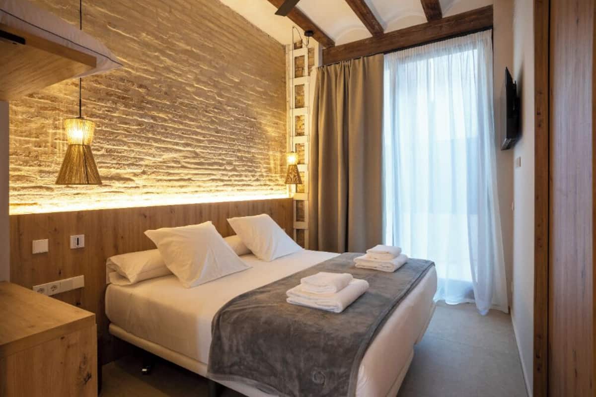 A hotel room with a wooden headboard, a large bed with white linens, a grey throw blanket, and a flat-screen TV mounted on the wall.