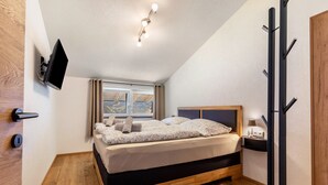 Un dormitorio con cama, televisor montado en la pared, ventana con cortinas y perchero.