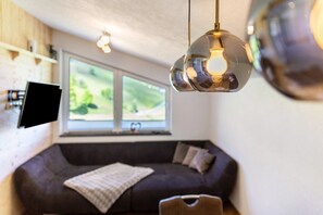 A modern living room with a dark sofa, a mounted TV, and a large window with a view of greenery. - Your Grubgut Getaway – Vacation with Mountain Views (Taxenbach)