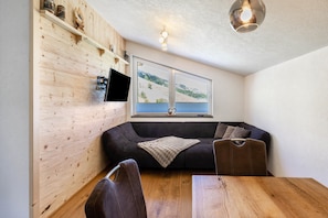 A modern living room with a wooden wall, a black sofa, a dining table, and a TV mounted on the wall. - Your Grubgut Getaway – Vacation with Mountain Views (Taxenbach)