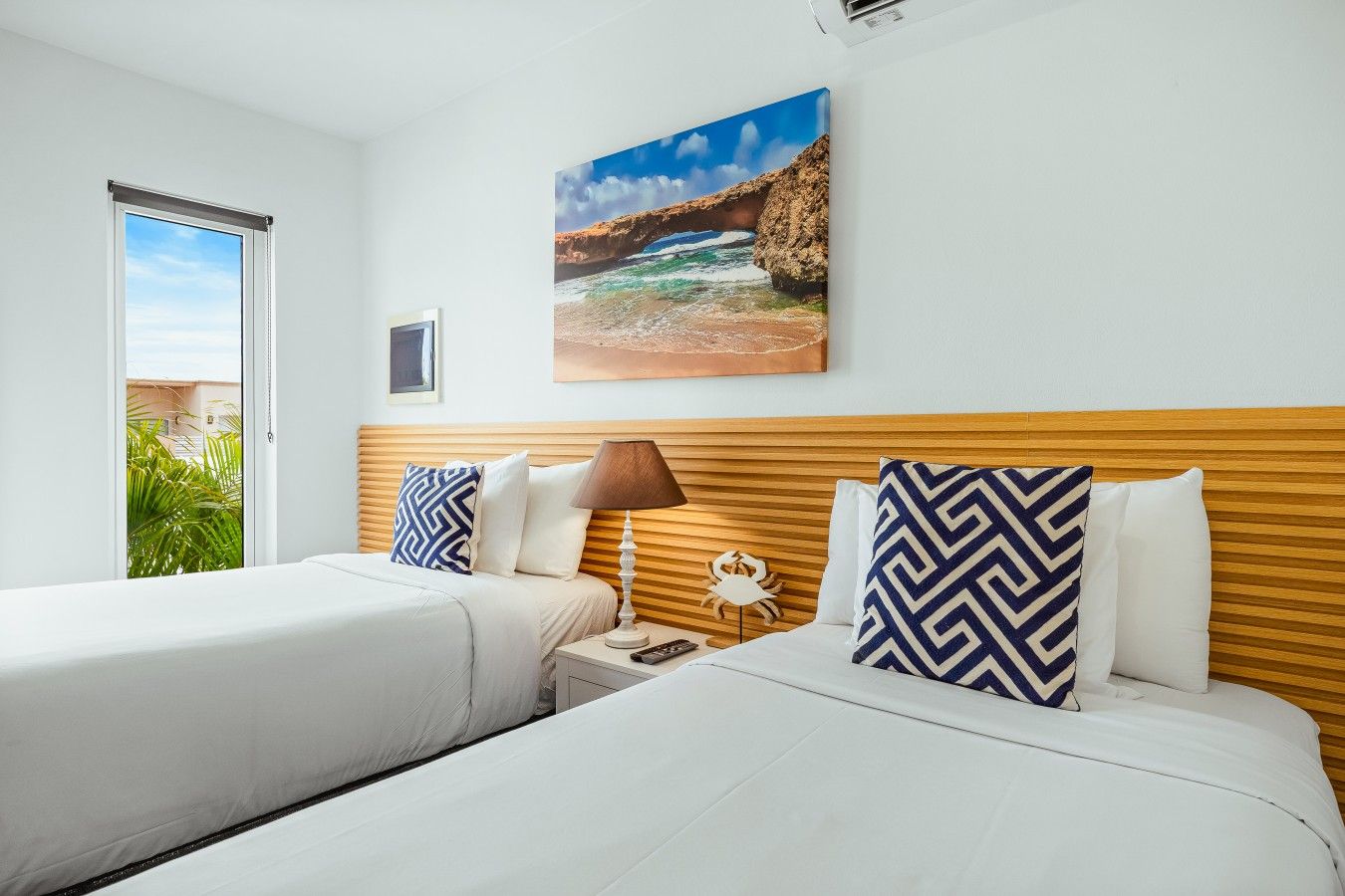 Une chambre d’hôtel avec deux lits, une tête de lit en bois, un tableau représentant un paysage côtier et une vue sur la verdure à l’extérieur.