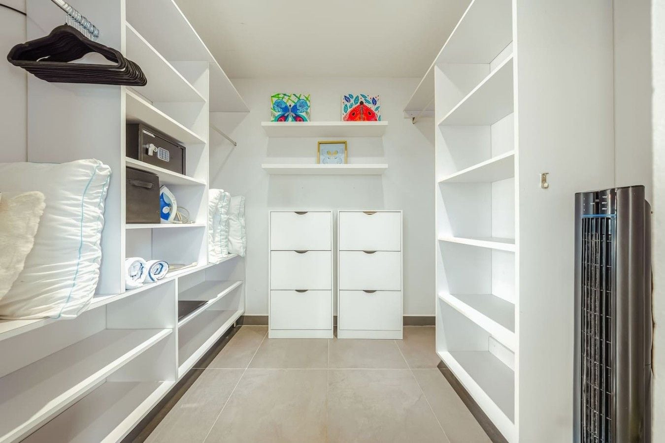A walk-in closet with white shelving, a central storage unit with drawers, and a black appliance on the right.