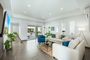 A modern living room with a sofa, armchairs, and a coffee table. There is a TV mounted on the wall, and a large window provides natural light. - Lux 3-Bedroom Villa with Private Pool by Bocobay (Noord)
