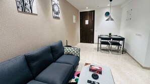 A living room with a dark grey sofa, a coffee table with remote controls and a box, a dining area with a table and chairs, and a wall with a clock and a framed picture.