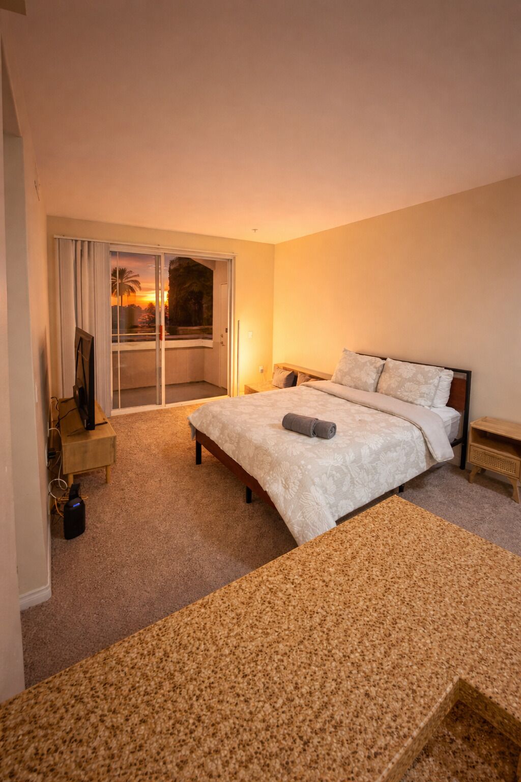 A bedroom with a bed, a desk, a chair, a TV, and a view of the sunset through the window.