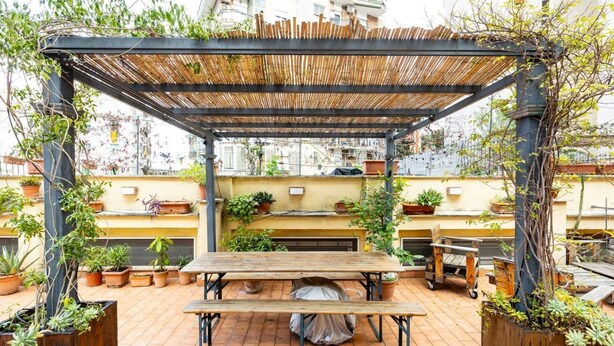 Um terraço no topo com uma mesa e bancos de madeira, rodeado por plantas em vasos e uma pérgula de bambu.