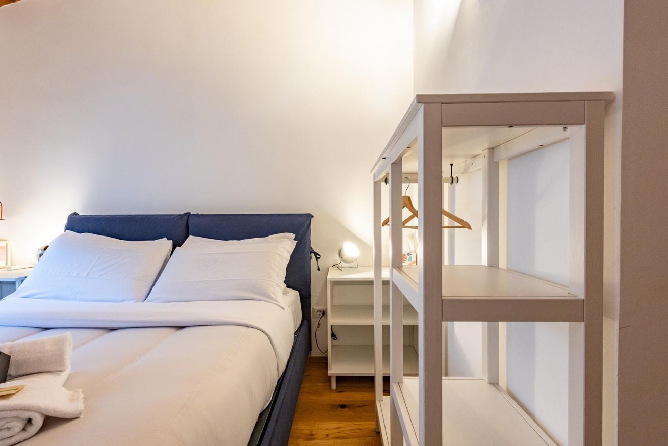A bedroom with a bed, white linens, a bedside table with a lamp, and a wardrobe with hanging space.