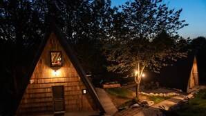 Eine dreieckige Holzhütte mit Tür und Fenster, umgeben von Bäumen und einem gepflasterten Weg.