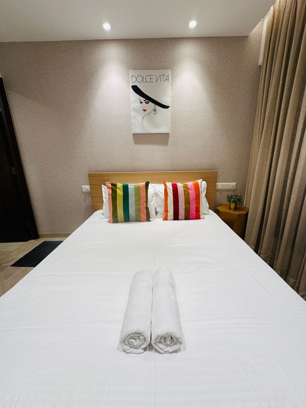 A neatly made bed with white linens and colorful striped pillows, a rolled white towel, and a wall-mounted artwork with the text "DOLCE VITA".