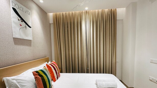 Hotelkamer met een bed, kleurrijke kussens, een wit hoofdeinde, een groot raam met gordijnen en een kunstwerk aan de muur.