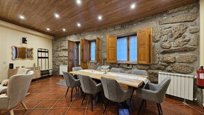 Un comedor con una larga mesa de madera, sillas grises tapizadas, paredes de piedra, contraventanas de madera y un extintor.