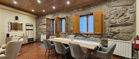 A dining area with a long wooden table, grey upholstered chairs, stone walls, wooden shutters, and a fire extinguisher.