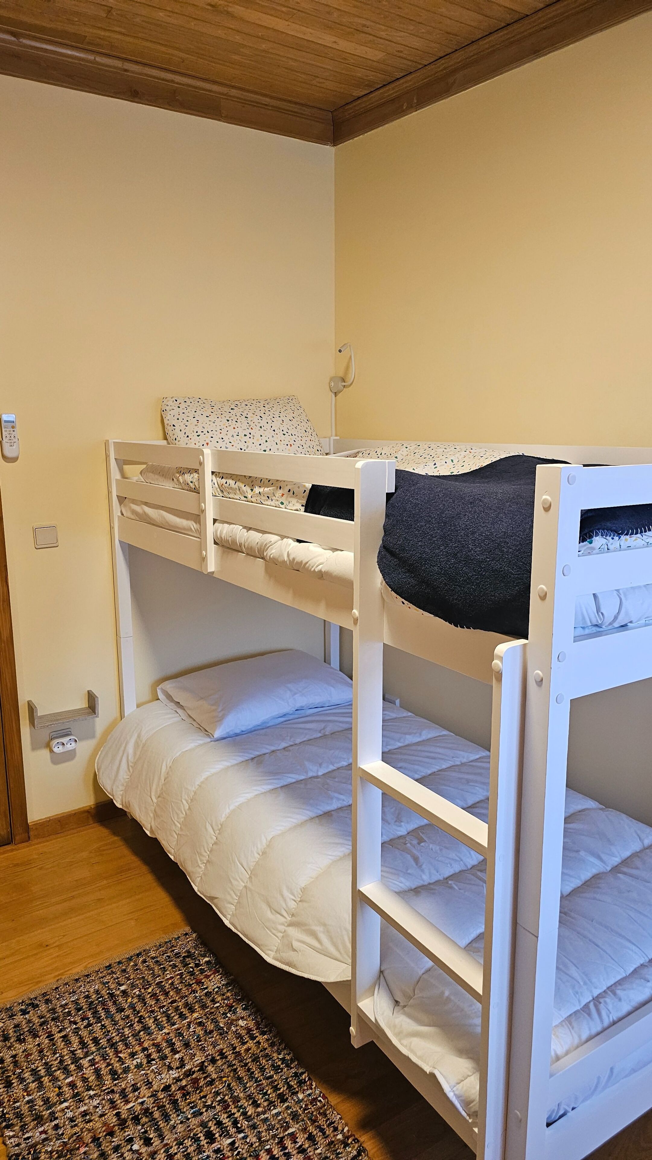 A bunk bed room with a wooden ceiling, a bedside table with a lamp, and a power outlet on the wall.