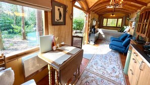 A cozy cabin interior with a wooden ceiling, a dining area with a laptop, a blue sofa, and a view of the forest through large windows.