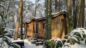 A wooden cabin with a porch and outdoor seating area, surrounded by snow-covered trees.