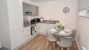 Une cuisine compacte avec des meubles blancs, un plan de travail noir et un coin repas comprenant une table en verre et des chaises grises.