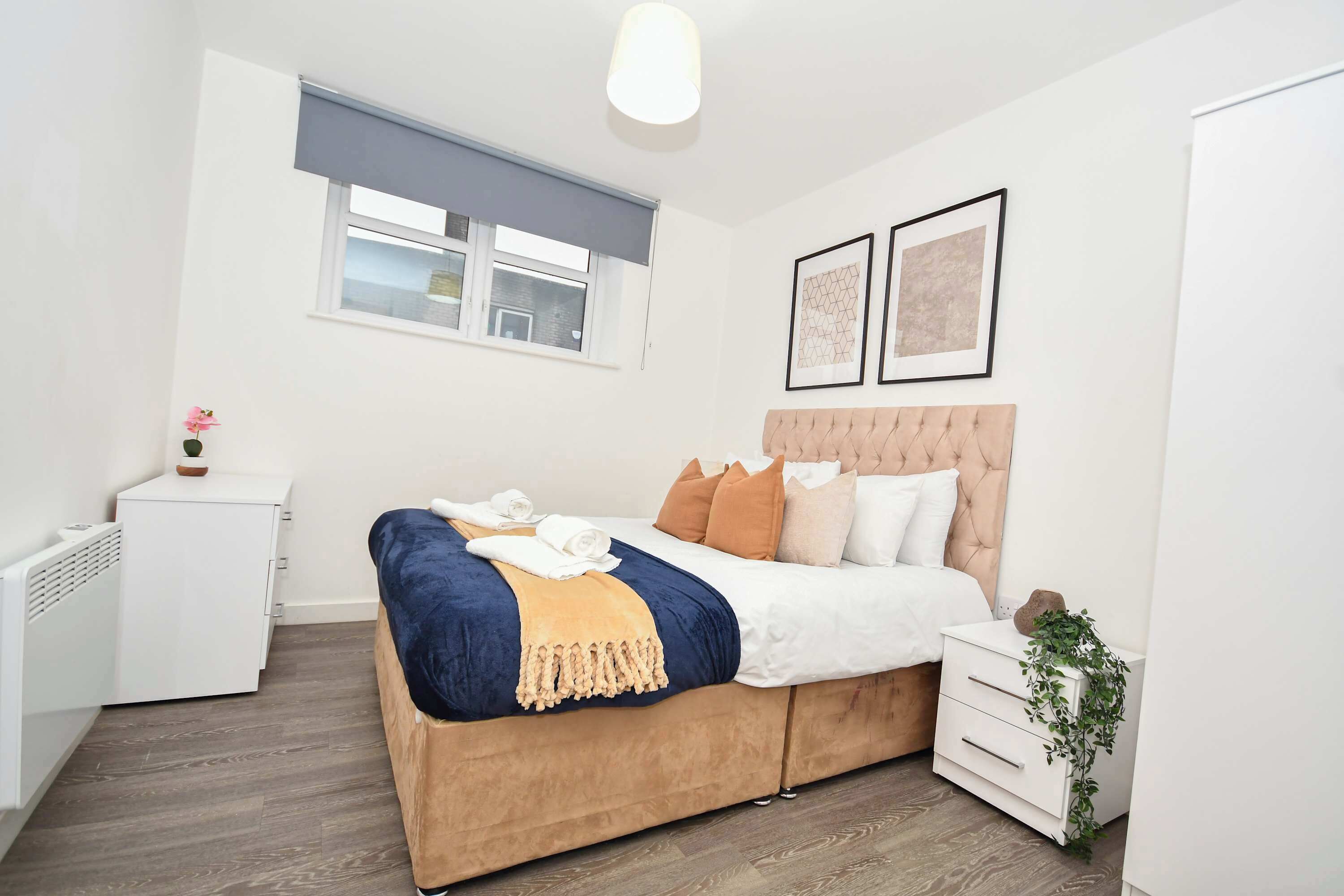 Een slaapkamer met een groot bed, een nachtkastje, een radiator, een raam met zonwering en twee ingekaderde foto’s aan de muur.