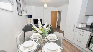Un espace repas moderne avec une table en verre pour quatre personnes, entourée d’armoires blanches et d’un canapé gris.