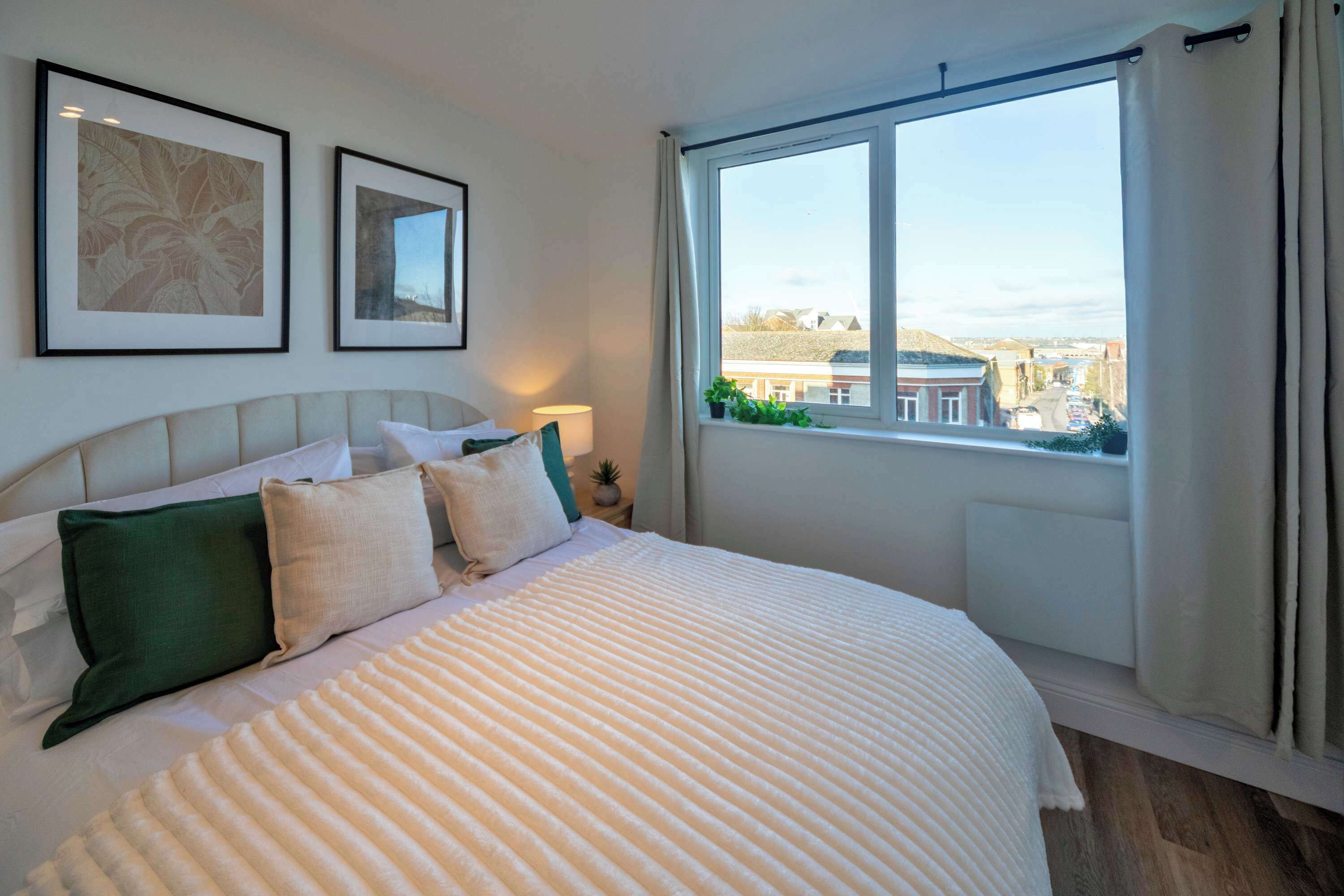 A neatly made bed with a striped blanket, a headboard, and two pillows. There are framed pictures on the wall, a window with curtains, and a view of buildings outside.