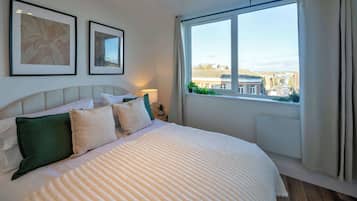 A neatly made bed with a striped blanket, a headboard, and two pillows. There are framed pictures on the wall, a window with curtains, and a view of buildings outside.