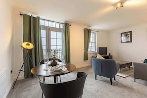 Una sala moderna con una mesa de comedor preparada para dos comensales, un sofá, un televisor y una lámpara de pie.