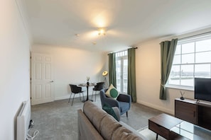 En stue med sofa, lænestol og et lille bord. Der er et vindue med gardiner, en dør og en radiator.