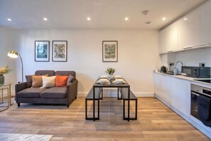 A modern living room with a sofa, a dining table set for four, and a kitchen area with appliances. - Spacious & Bright 1 Bedroom Apartment in Gravesend (Gravesend)