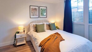 Ein ordentlich bezogenes Bett mit weißer Decke und brauner Tagesdecke, an beiden Seiten zwei Nachttische mit Lampen und einer kleinen Pflanze.