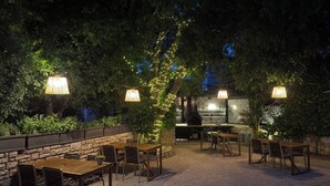 Eine Außenpatio mit Steinmauern, Holztischen und -stühlen, umgeben von Bäumen und beleuchtet durch hängende Lampen.