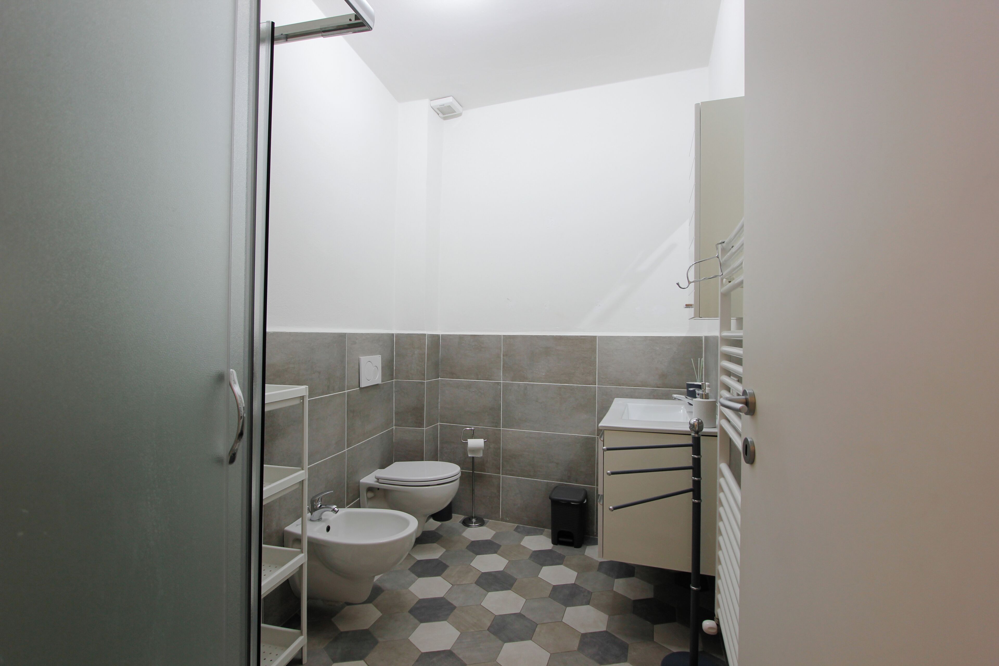 A compact bathroom with a toilet, bidet, and sink, featuring hexagonal patterned tiles and a glass shower enclosure.