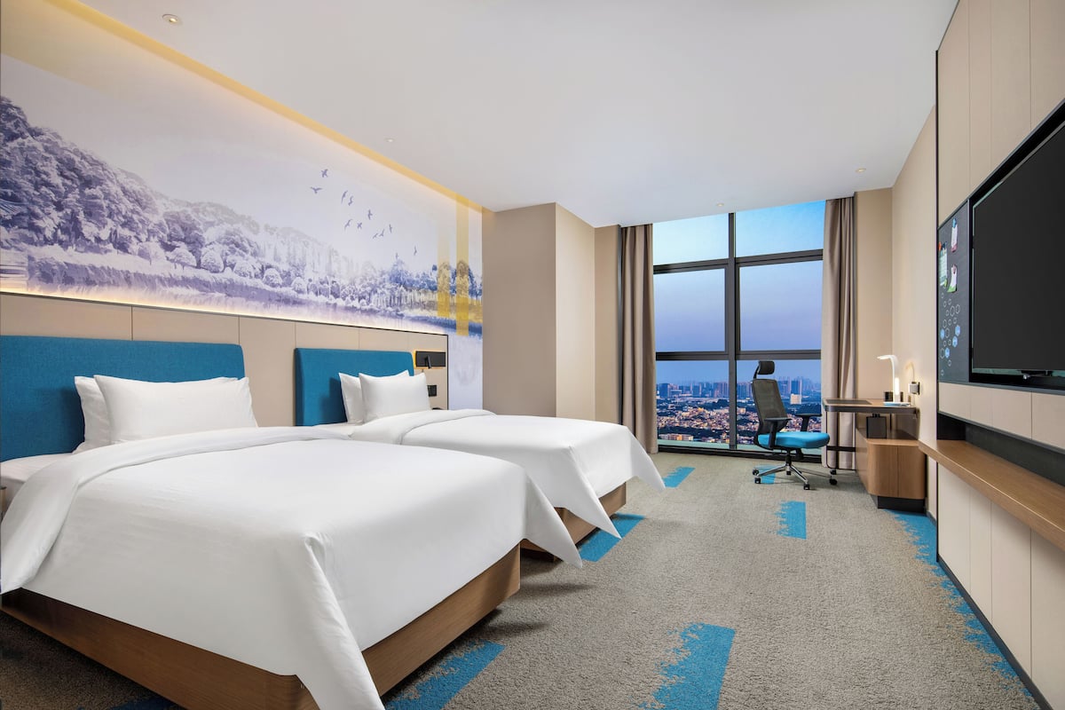 Une chambre d’hôtel avec deux lits, un bureau, une télévision et une grande fenêtre donnant sur la ville.