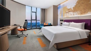 A hotel room with a large bed, a desk with a television, a chair, and a view of the city through large windows. - Hampton by Hilton (Pingxiang)