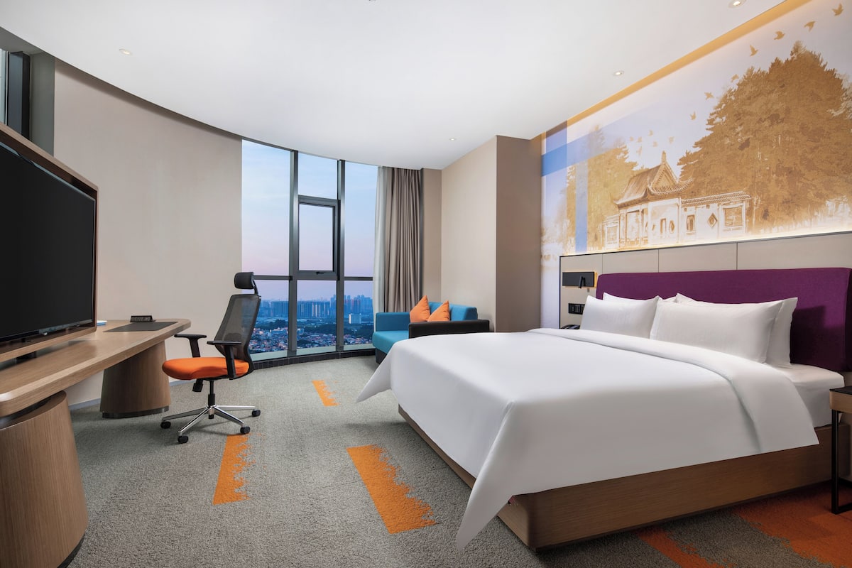 Une chambre d’hôtel avec un grand lit, un bureau avec une télévision, une chaise et une vue sur la ville grâce à de grandes fenêtres.