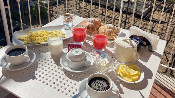 Een balkon tafel met ontbijtartikelen, zoals roerei, croissants, sap, koffie en een fruitschaal.