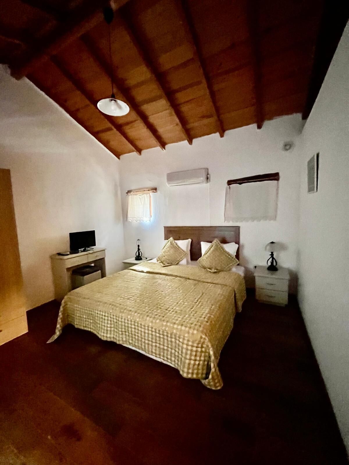 Un dormitorio con techo de madera, una cama con colcha a cuadros, dos mesitas de noche con lámparas, un aire acondicionado instalado en la pared y una ventana con cortina.