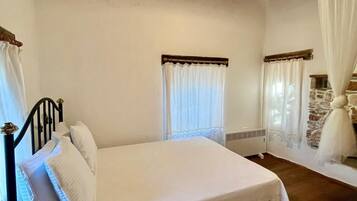A bedroom with a bed, a nightstand, a window with curtains, a stone wall, and a wooden ceiling.