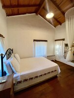 A bedroom with a bed, a nightstand, a window with curtains, a stone wall, and a wooden ceiling.