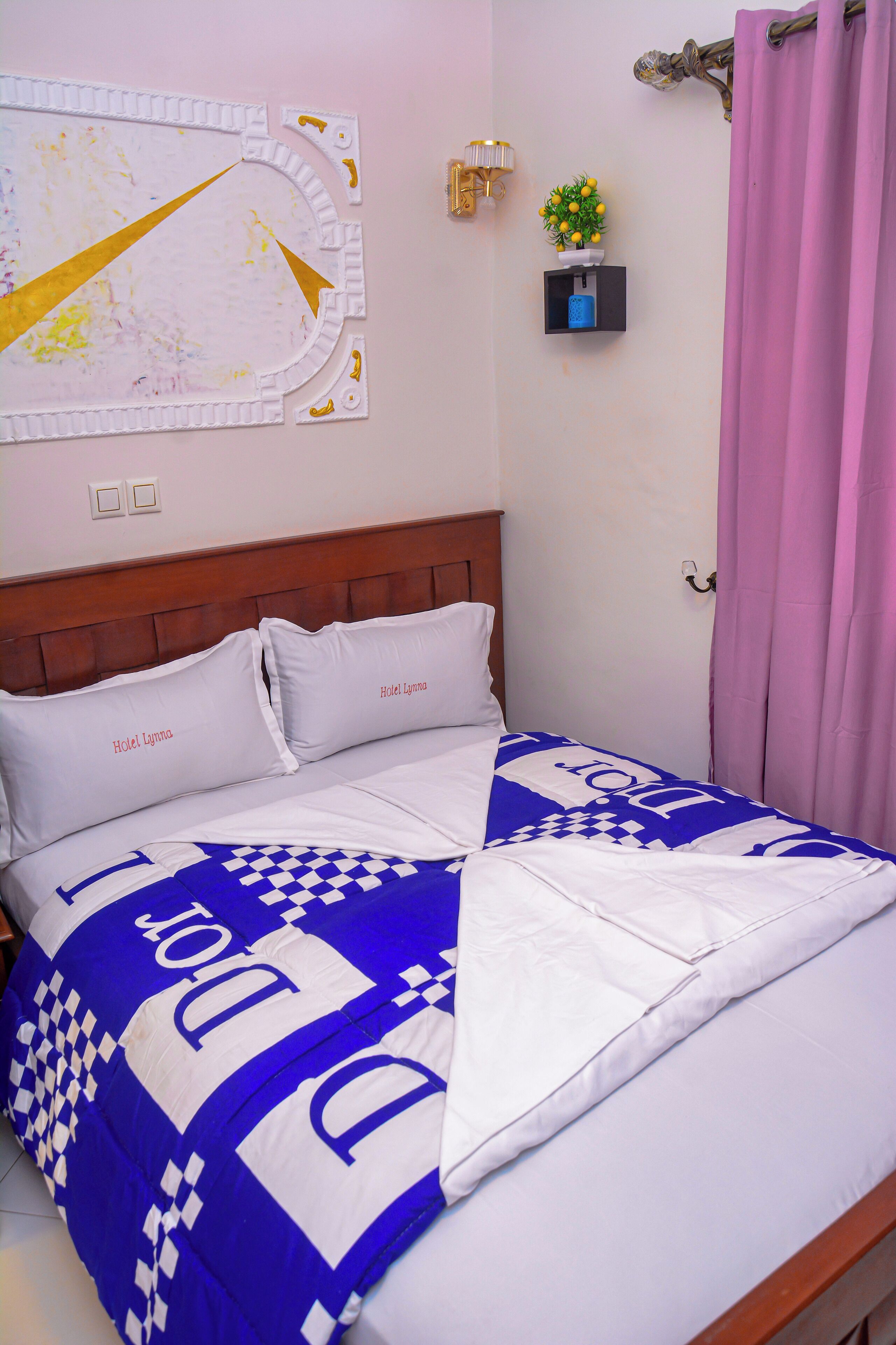 Una habitación de hotel con una cama, almohadas con la etiqueta 'Hotel Lygna', un estante en la pared con una planta y una cortina rosa.