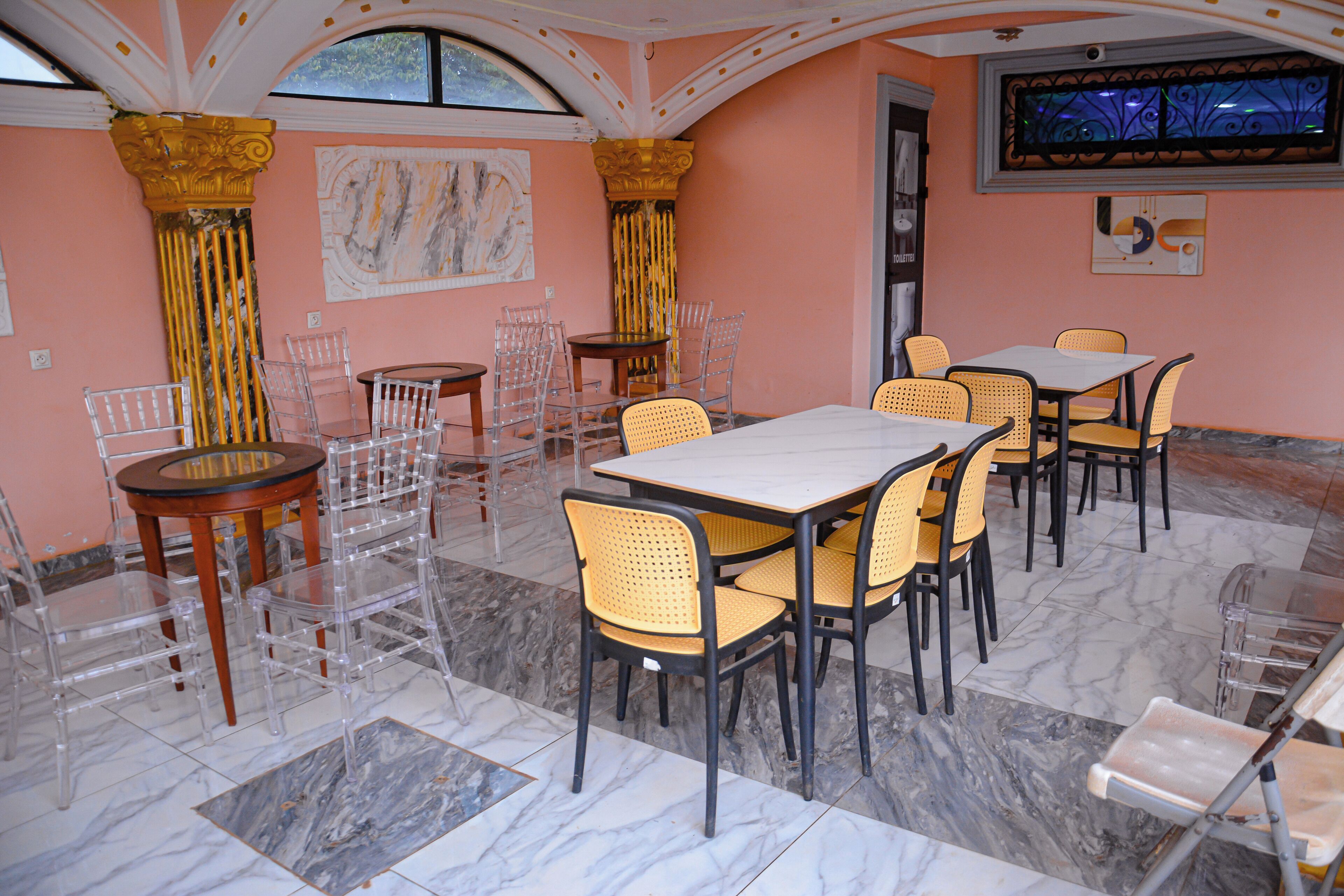 Comedor con suelo de mármol, sillas transparentes y mesas, con un ventanal grande y columnas decorativas.