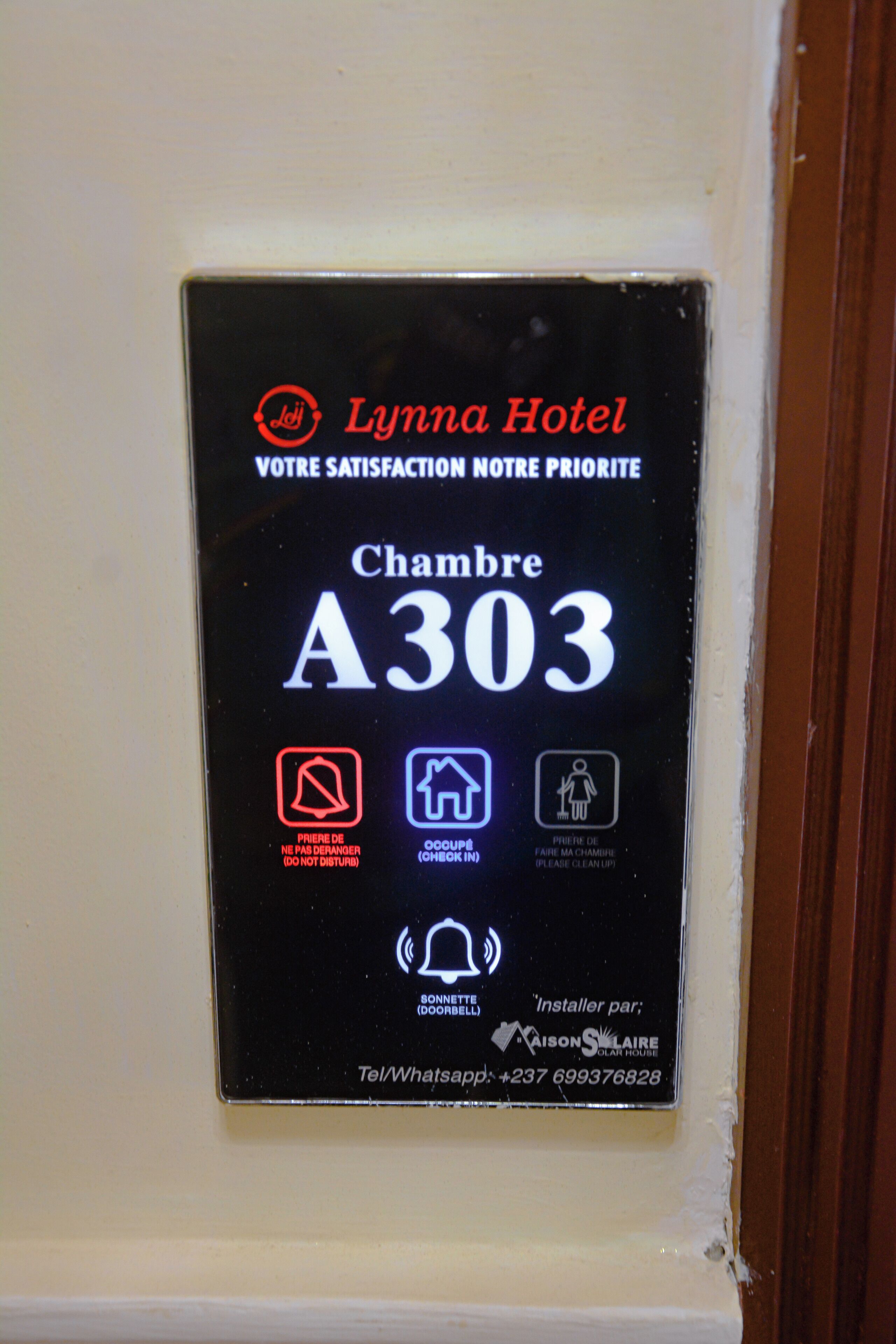 A hotel room number sign for Lynda Hotel, room A303, with icons for a do not disturb sign, a house icon, and a bell icon.
