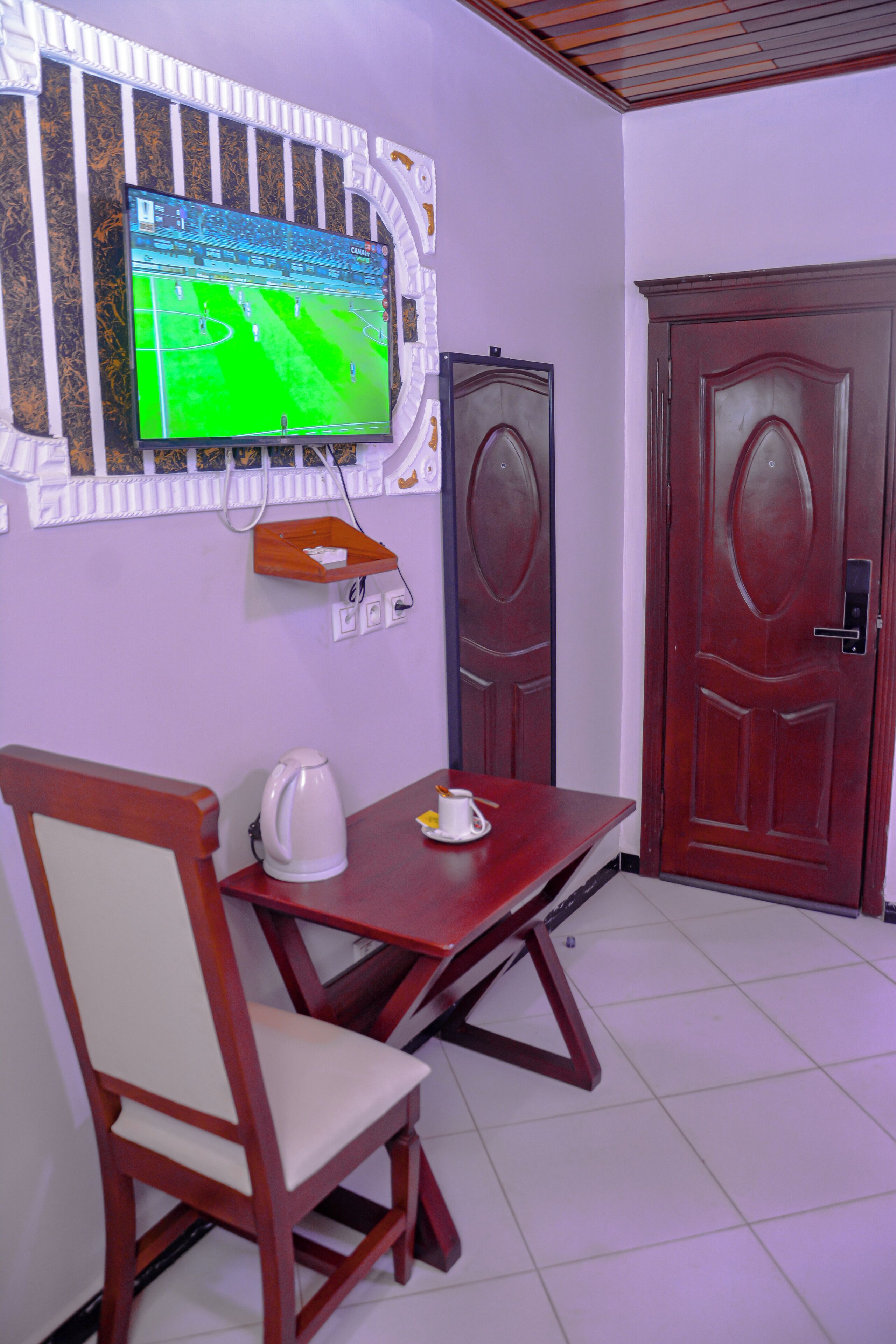 A room with a TV mounted on the wall, a wooden table with a chair, a door, and a small shelf with items.