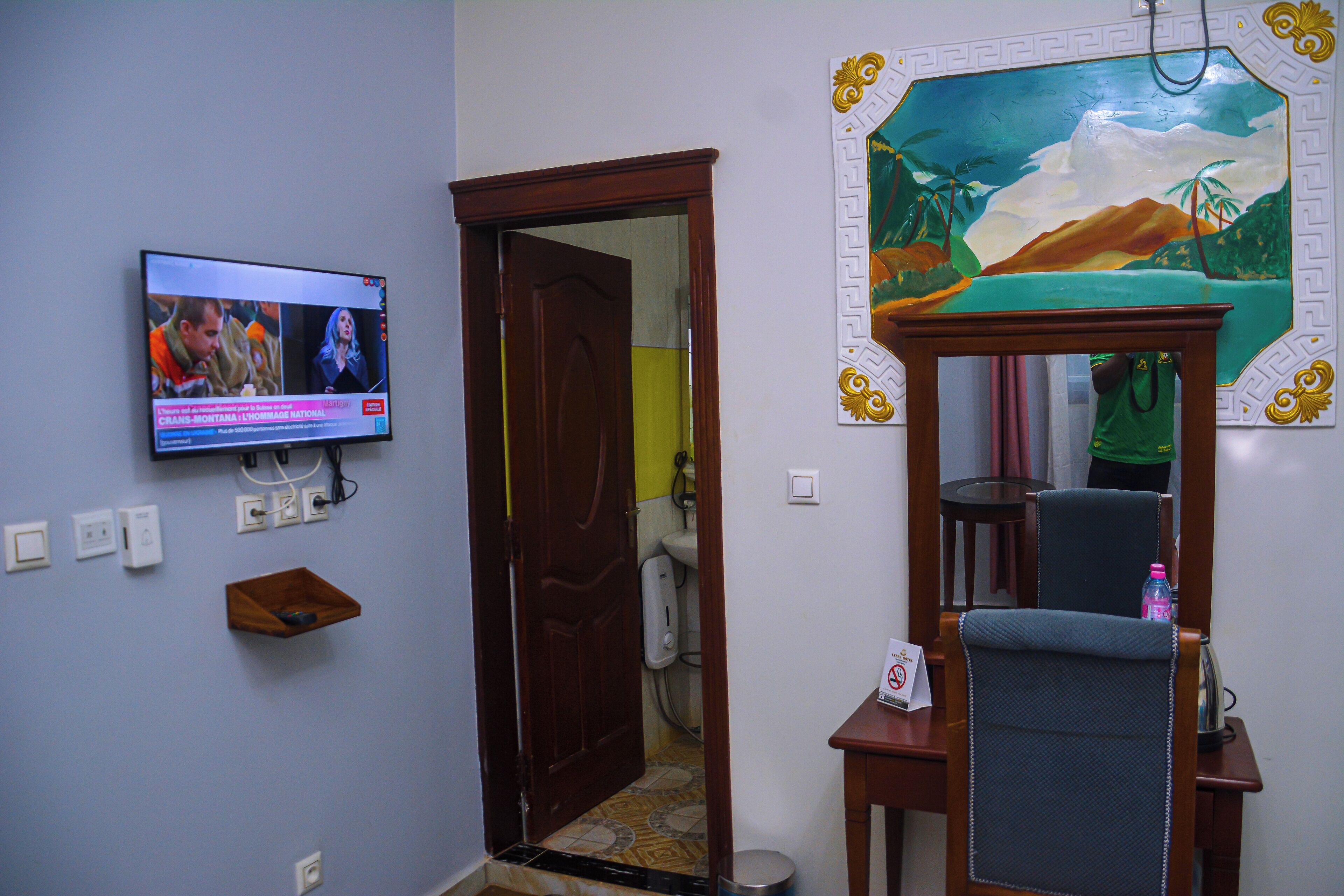 A room with a television mounted on the wall, a wooden door, a mirror with a decorative frame, and a small shelf.