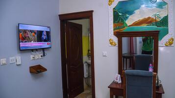 A room with a television mounted on the wall, a wooden door, a mirror with a decorative frame, and a small shelf.