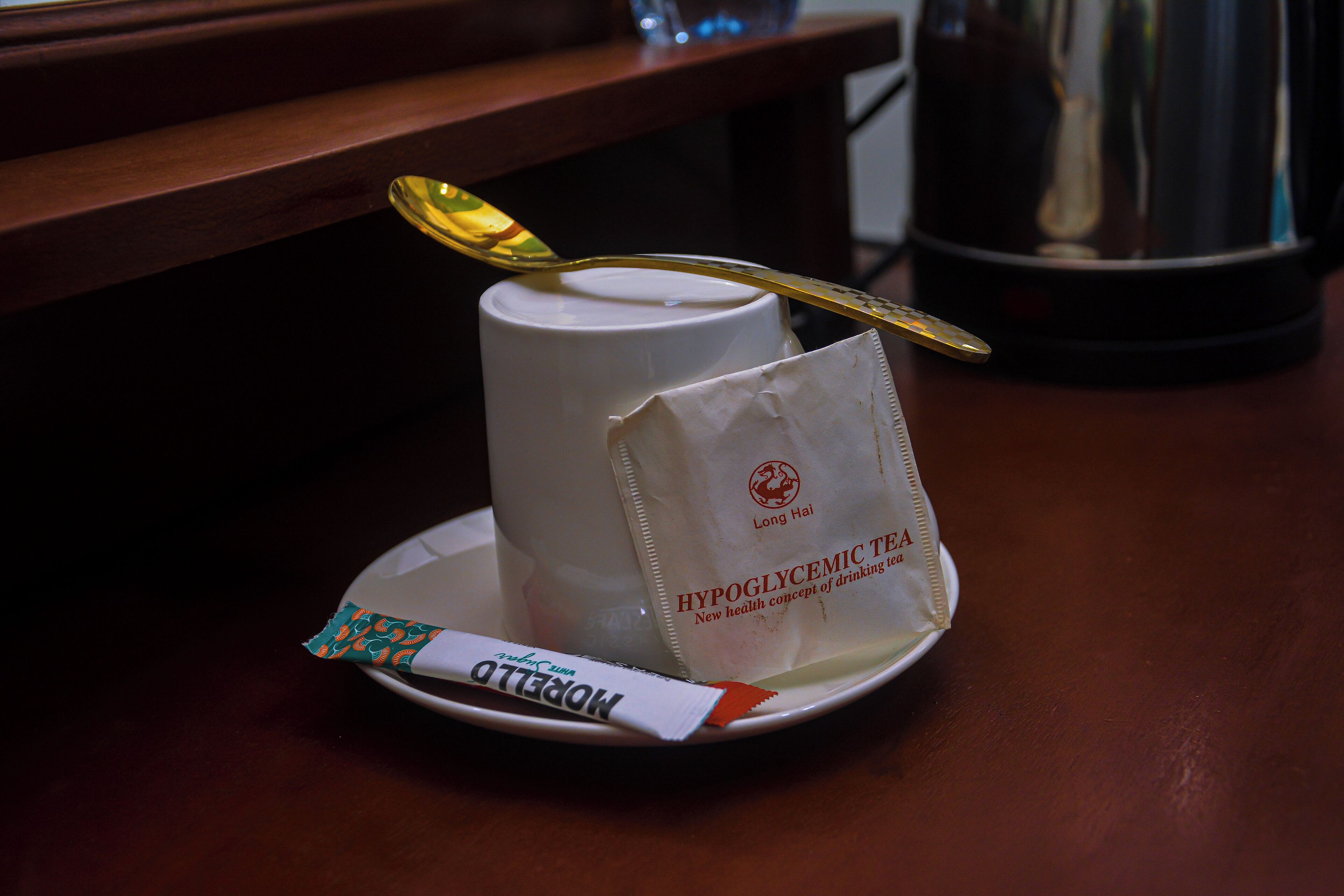 A white cup with a spoon on a saucer, a packet of 'Hypoglycemic Tea' on a plate, and a metallic object in the background.